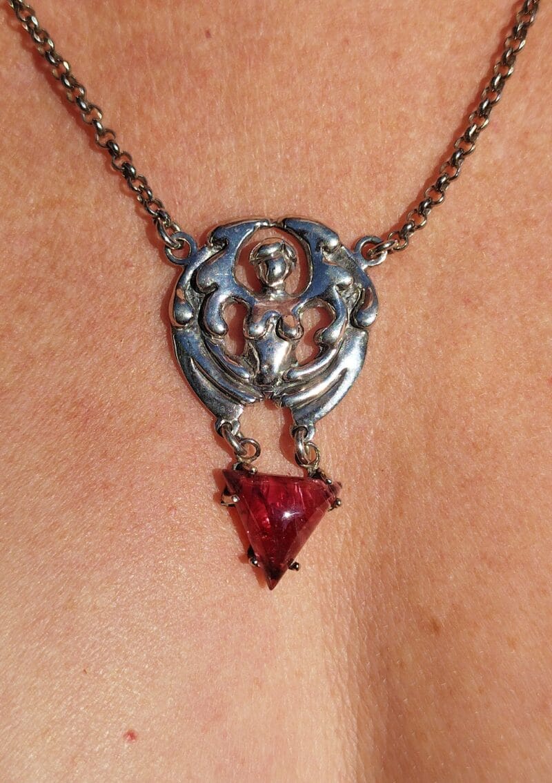 Zilveren ketting met een hanger in de vorm van een beschermengel. Onder de engel hangt een rode, driehoekige spinel.