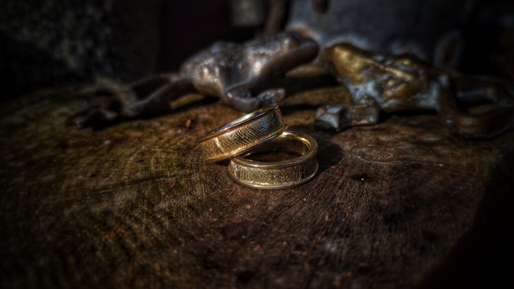 Een set unieke, gouden trouwringen waarin de vingerafdrukken van het koppel zijn verwerkt. De ringen liggen op een rustieke houten schijf waarop de jaarringen duidelijk zichtbaar zijn.