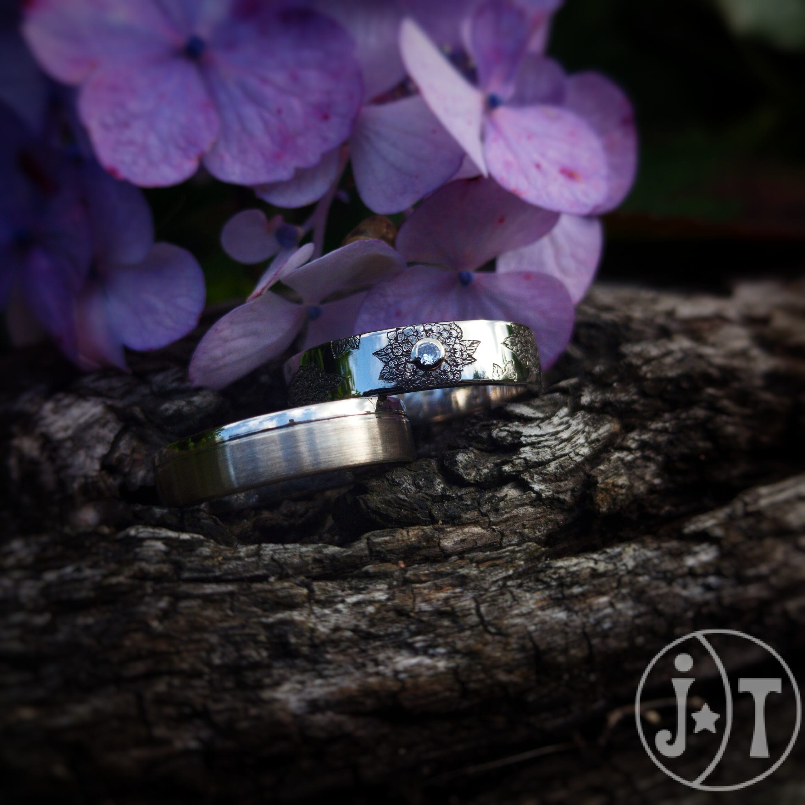 Witgouden trouwringen op een stuk boomschors met paarse hedera bloemen. De herenring heeft een matte afwerking en gepolijste randen. De damesring is gegraveerd met een hedera bloemen met in het midden een diamant. trouwringen-goudsmid-drenthe